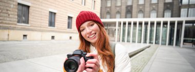 Beautiful readhead girl, photographer with professional camera takes pictures outdoors, walking around city and taking photos, sightseeing.