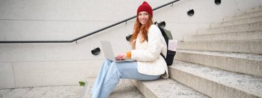 Portrait of young girl traveller, sitting with backpack and map of city, working on laptop, connecting to public wifi and sitting on stairs outdoors, using internet to book hotel room.