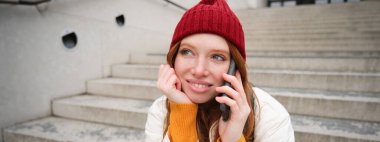 Beautiful smiling redhead female model, sits on street and talks on mobile phone, uses smartphone app to call abroad, laughing during telephone conversation.