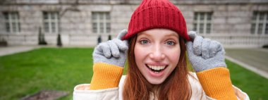 Cute girl student in red hat, warm gloves, sits in park, smiles and looks happy. Copy space