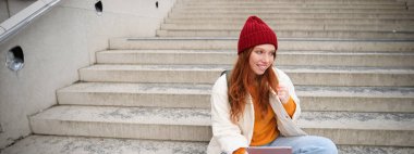 Young beautiful girl with long red hair, smiles, uses internet application, connect to wifi near campus, sits on stairs with digital tablet.