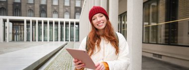 Stylish ginger girl, tourist walks with digital tablet around city, woman connects to iternet on her gadget, looking up information, texting message.