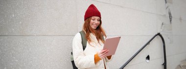 Young redhead woman with red hat, uses her digital tablet outdoors, stands on street with gadget, connects to wifi internet and searches for a location in internet.