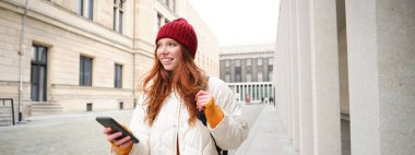 Beautiful smiling girl, tourist with backpack, holding smartphone, using map on mobile phone application, looking for sightseeing in internet app, standing outdoors.