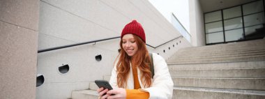 Redhead girl in red hat, sits on stairs and uses mobile phone. Modern woman holding smartphone, texting message, using telephone application outdoors.