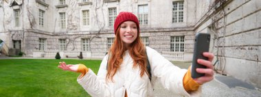 Portrait of happy tourist, girl with backpack, video chats, shows building, sightseeing attraction to friend, poses near landmark and smiles.