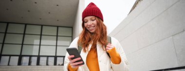 Happy girl student in red hat, holds smartphone, tourist looks at map app on her phone, explores sightseeing, texts message, looks for couchsurfing, rents place to stay online.