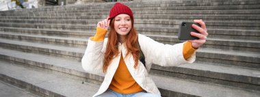 Stylish young girl in red hat, takes photos on smartphone camera, makes selfie as she sits on stairs near museum, posing for photo with app filter.