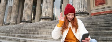 Young redhead woman sits on stairs outdoors and waves hand at smartphone camera, video chats with friends, connects to public wifi.