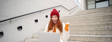 Smiling redhead girl, young woman typing on laptop keyboard, sitting outdoors on stairs with computer, working remote, doing her homework on fresh air.