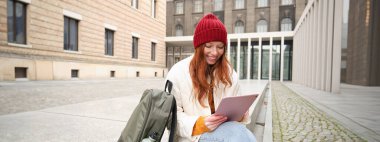 Redhead girl smiles, sits outdoors near building with digital tablet, thermos and backpack, connects to public internet and searches smth online on her gadget.