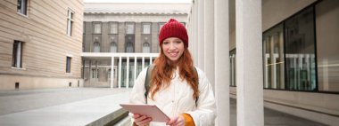 Happy redhead girl in red hat, walks around city with digital tablet, connects to public internet wifi and looks for route, looks at map on her gadget.