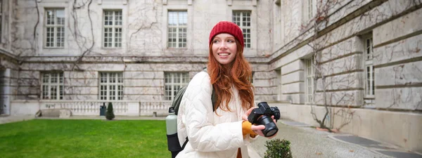 stock image Happy redhead girl tourist, takes photos, photographer with professional camera walks around city and captures beautiful pictures.