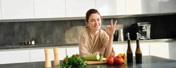 Portrait of smiling brunette girl making vegan dinner, showing okay sign, recommending healthy food, chopping vegetables in kitchen for salad or for vegan breakfast.