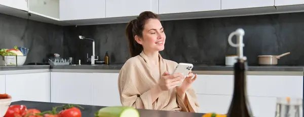 stock image Portrait of beautiful young woman with smartphone, sitting in the kitchen, cooking dinner, vegetables on chopping board. Girl watching cooking recipe on mobile phone.