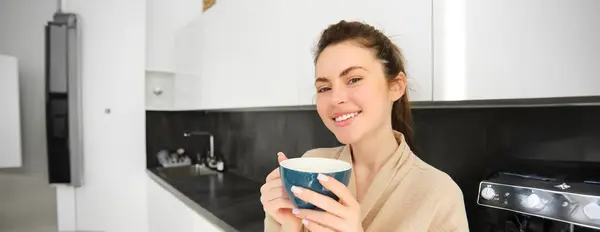 stock image Portrait of happy, smiling modern woman, starts her day with morning mug of tea. Girl drinking coffee in the kitchen, standing with mug and looking relaxed.