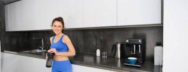 Mutfakta duran ve spor şişesinden su içen mavi etek giyen genç bir kadın portresi..