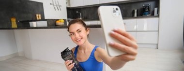 Refah ve spor. Güzel genç bir kadın akıllı telefondan selfie çekiyor, evden fitness eğitimi alıyor, su içiyor, susuz kalmıyor ve egzersiz yapıyor..