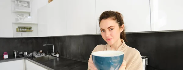 stock image Portrait of good-looking young woman gives you cup of coffee, offering mug to you, standing in the kitchen, wearing bathrobe.