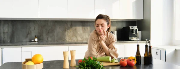 Portrait of woman thinks what to cook with vegetables, looking at chopping board with tomatoes with thoughtful face, standing in kitchen preparing food.