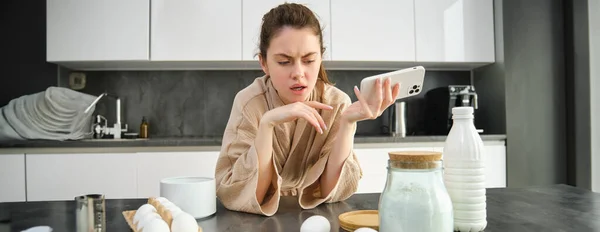 stock image Attractive young cheerful girl baking at the kitchen, making dough, holding recipe book, having ideas.