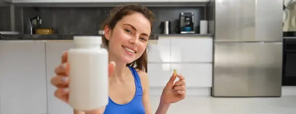 Close up portrait of fitness woman, girl workout from home, showing bottle of vitamins, dietary supplement pills, recommending buds.