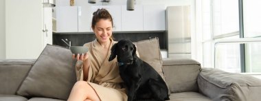 Bornozlu mutlu genç bir kadın, evde köpeğiyle vakit geçirmeyi seviyor, mısır gevreği yiyor, yavru köpekle kahvaltı ediyor, kanepede oturuyor..