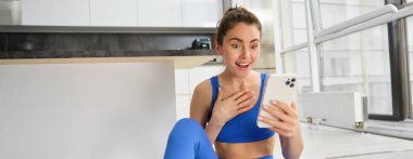 Şaşırmış, mutlu, genç bir sporcunun gülümseme ve heyecanla akıllı telefona bakması, telefon mesajında harika haberler okuması, mavi spor ayakkabılar ve taytlarla oturması..