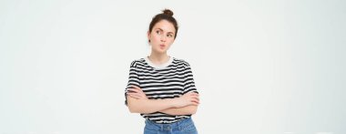 Düşünen, düşünceli, düşünceli dudakları büzüşmüş, kolları göğsünde beyaz arka planda duran genç bir kadın tasviri..