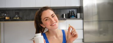 Gülümseyen, sağlıklı genç bir kadının portresi, elinde tabletler, evde spor yaptıktan sonra vitamin takviyesi almak..