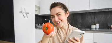 Gülümseyen güzel bir kadın resmi, mutfakta oturuyor, akıllı telefon ve domates tutuyor, mutlu görünüyor, evine yiyecek siparişi veriyor, cep telefonu uygulaması kullanıyor, tarifi için çevrimiçi sebze alıyor..