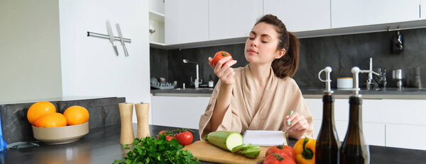Portrait of beautiful smiling woman, writing her healthy menu, eating tomato while cooking, making grocery list, sitting in the kitchen.
