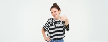 Kendine güveni olan, rahat giyinen, kamerayı işaret eden, gülen ve gülümseyen, beyaz arka planda duran genç bir kadın..