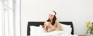 Güzel bir kadının portresi, uyku tulumu ve pijama giymiş, yatakta kahve içen, aydınlık ve rahatlatıcı sabahının tadını çıkaran..
