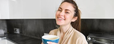 Gülümseyen kadının portresi kahve içer, mutfakta durur ve sabahları lezzetli bir fincan kapuçino içer, kameraya mutlu bakar..