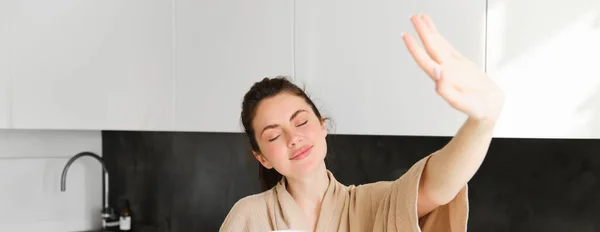 stock image Portrait of beautiful, happy young woman covers her eyes from morning sunlight, waking up and drinking coffee, holding mug, posing in the kitchen.