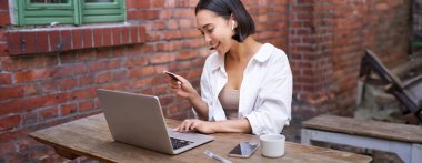 Gülümseyen Asyalı kadın dizüstü bilgisayarla oturuyor, online alışveriş için kredi kartıyla ödeme yapıyor, banka hesap bilgilerini gönderiyor, kafede oturuyor..