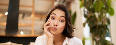 Kafede oturmuş, neşeli bir şekilde sohbet eden, kameraya gülümseyen komik Asyalı genç kadının yakın portresi..