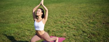 Asyalı genç bir kadının portresi mutlu bir şekilde, yoga yaparken vücudunu esnetiyor, mindere basıyor ve parkta egzersiz yapıyor..