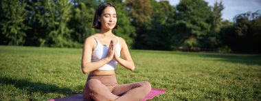 Parkta çimenlikte meditasyon yapan bir kadın, spor minderinde oturuyor, rahatlıyor, temiz hava soluyor..