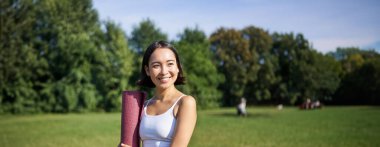 Genç Asyalı fitness kızının dikey portresi yoga için lastik minderle yürüyor, parkta temiz hava eğitimi alıyor, spor kıyafetleri giyiyor..