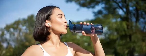 Happy asian sportswoman, runner drinks water from bottle while running, workout on fresh air in park.