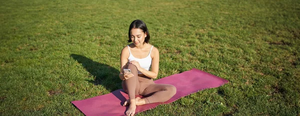 Woman in park, watching yoga video on smartphone, meditating on fresh air, sitting on rubber mat.