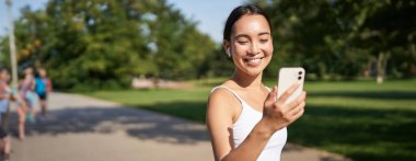Mutlu Asyalı fitness kızı koşuyor ve istatistiklerini kontrol ediyor. Akıllı telefon uygulamasındaki günlük hedefler. Genç kadın koşuyor ve seyyar ekrana bakıyor.
