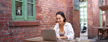 Mutlu Asyalı kız dizüstü bilgisayarda video izliyor, dışarıda ödevlerini yapıyor, birlikte çalışıyor, gülümsüyor ve çalışıyor, kahve içiyor..