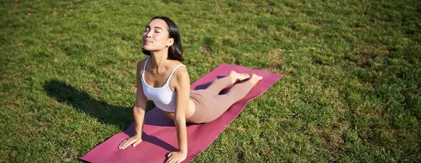 Smiling young sportswoman stretches on rubber mat in park, does yoga asana exercises, workout on fresh air in fitness clothing.