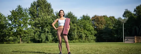 stock image Confident and sporty young asian woman, standing in park with rubber mat, wearing sportswoman, smiling at camera, workout on fresh air.