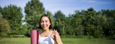 Heyecanlı genç bir kadın spor minderiyle ayakta duruyor, yoga kıyafetleri, iyi bir işaret, parkta egzersiz, dışarıda iyi bir eğitim..