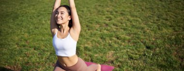 Asyalı genç bir kadının portresi mutlu bir şekilde, yoga yaparken vücudunu esnetiyor, mindere basıyor ve parkta egzersiz yapıyor..