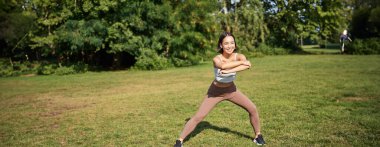 Genç Asyalı fitness kızı geriniyor, parkta egzersiz yapıyor, egzersiz yapıyor, çömelme hareketleri yapıyor..